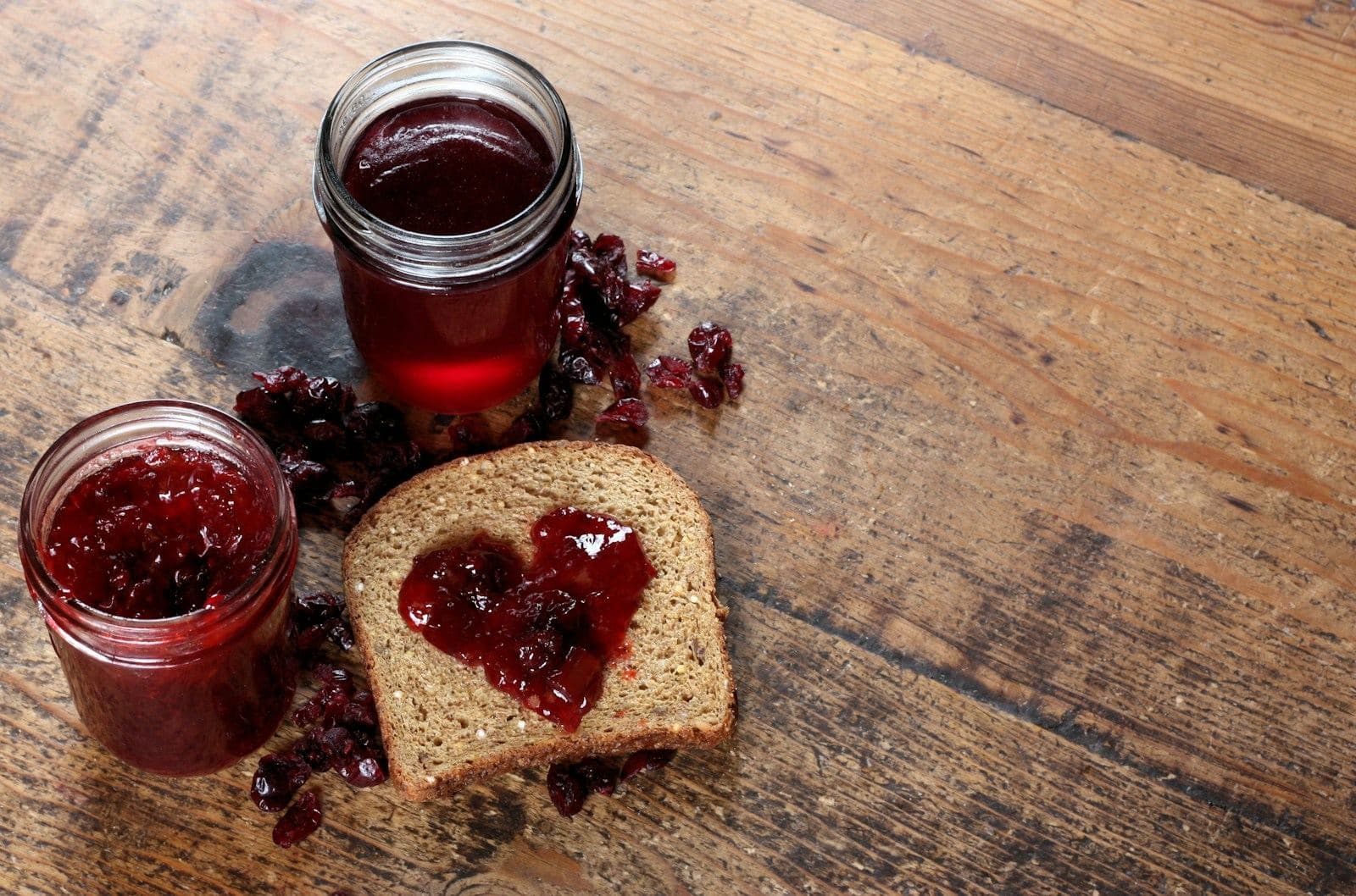 Two jars of red jam beside a slice of bread with a jam heart on a wooden table